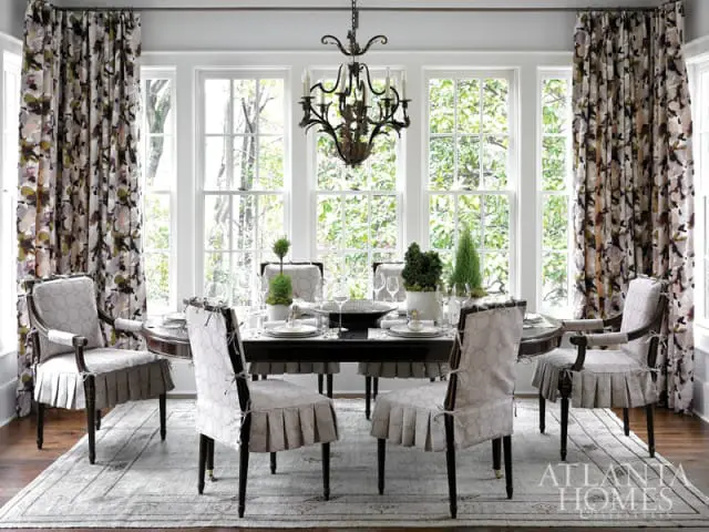 Dining room with dark wood table and chairs upholstered in patterned fabric, floor-to-ceiling windows with floral curtains, and a large patterned rug.