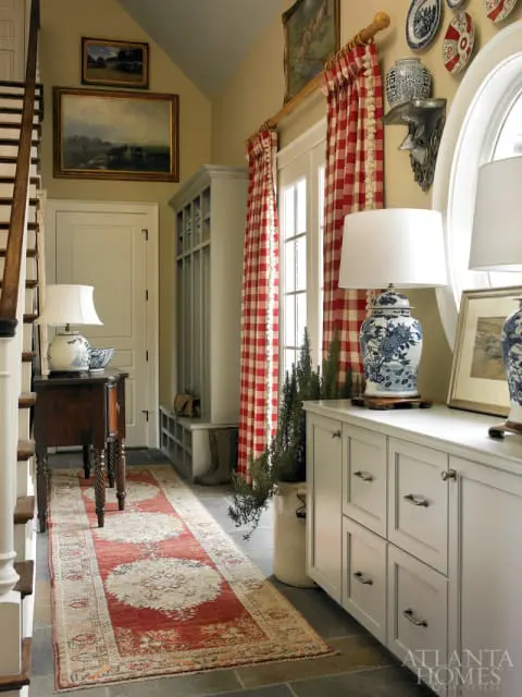 Hallway with a red and cream patterned runner rug, a wooden console table with a lamp, a gray built-in bench, and white cabinetry with decorative blue and white ginger jars. Red gingham curtains hang by a door.