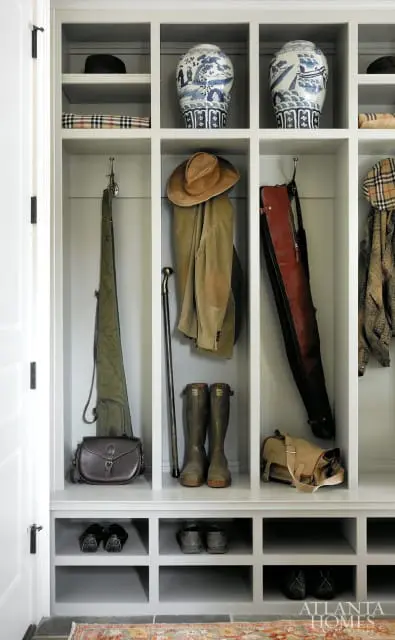 Built-in entryway storage with cubbies holding two blue and white ginger jars, a tan hat, a corduroy jacket, boots, bags, and shoes. A cane and gun case hang on hooks.