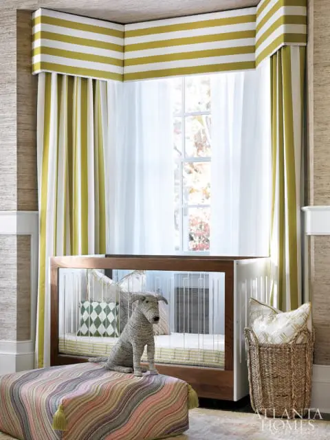 Nursery with a modern wooden crib, striped green and white curtains, a patterned ottoman with a stuffed dog, and a woven basket with pillows.