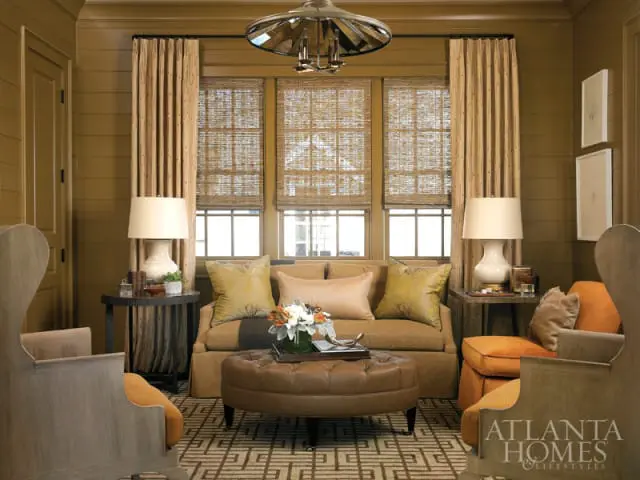 Living room with olive green paneled walls, featuring a tan sofa, two orange accent chairs, a brown leather ottoman, and bamboo window shades.