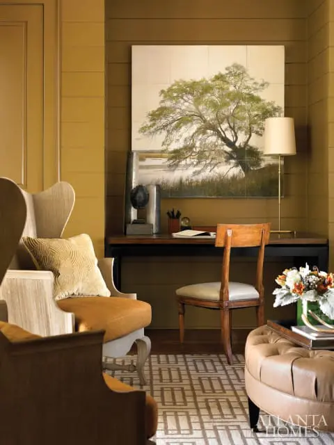 Study with mustard yellow paneled walls, a large painting of a tree over a desk, wingback chairs, a wooden desk chair, and a tufted leather ottoman.