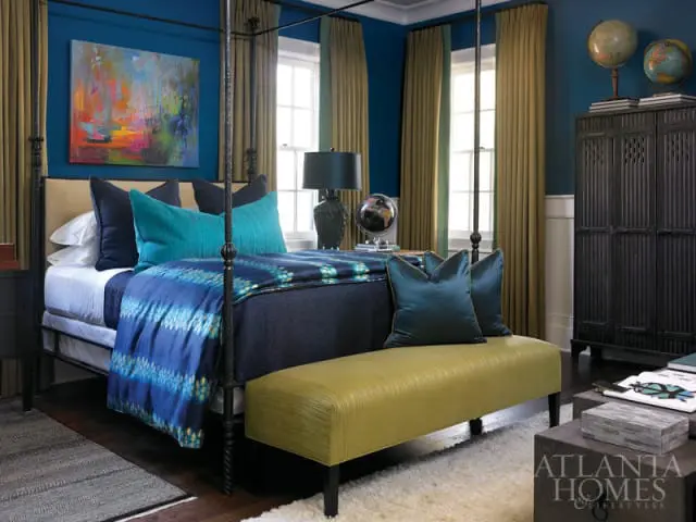 Bedroom with a dark blue wall, four-poster bed with navy and teal bedding, abstract art, and a yellow bench.