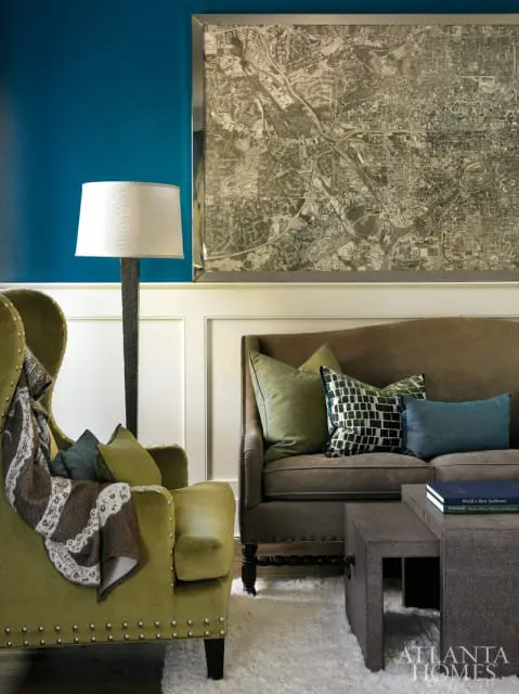 Living room with teal walls, a large framed map, a green wingback chair with a blanket, a brown sofa with green and blue pillows, and a textured coffee table.