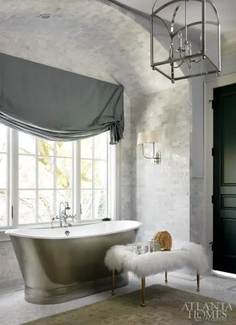 Luxury bathroom with a freestanding silver bathtub, marble tile walls and ceiling, a large window with a grey Roman shade, and a white fur bench with gold legs.