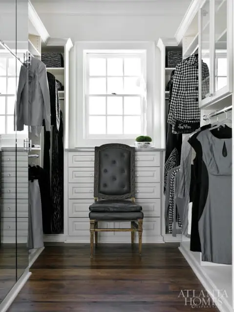 Walk-in closet with white built-in cabinetry, dark wood floors, and a tufted black leather chair in front of a window.
