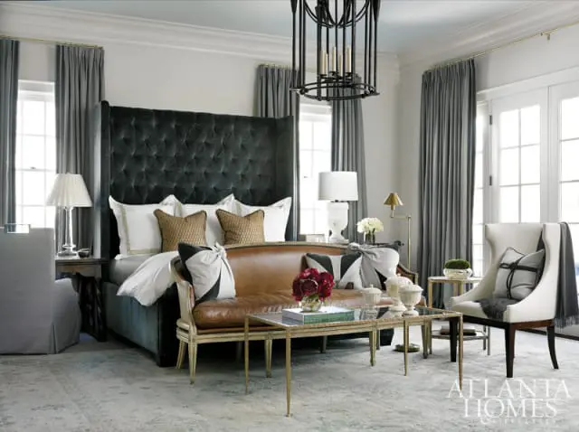 Bedroom with large tufted dark green headboard, leather sofa, wingback chair, and glass coffee table. Gray curtains frame windows and French doors.