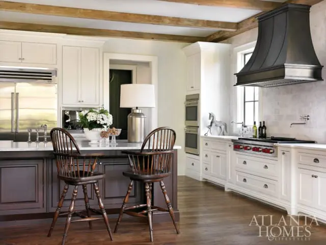 White kitchen with dark wood island, two wooden Windsor bar stools, stainless steel appliances, and a black metal range hood over a gas stove.