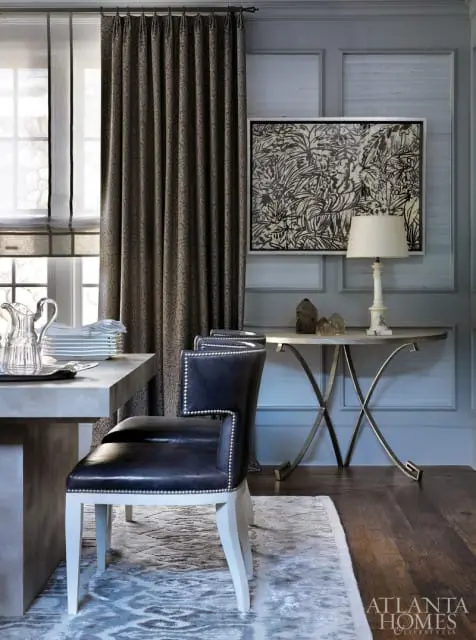 Dining room with a concrete table, dark leather chairs with silver nailhead trim, a console table with a lamp and artwork, and patterned curtains.