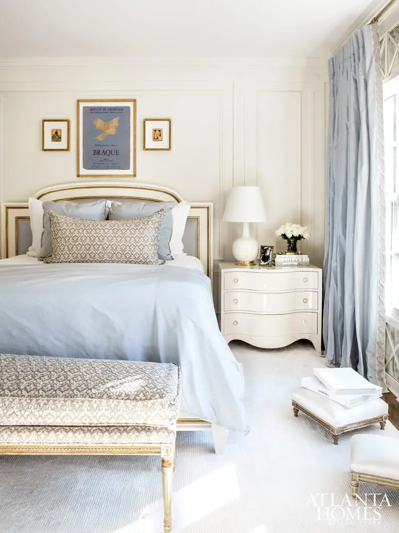 Bedroom with a light blue duvet, patterned accent pillow, and a tufted bench at the foot of the bed. A white nightstand holds a lamp and floral arrangement.