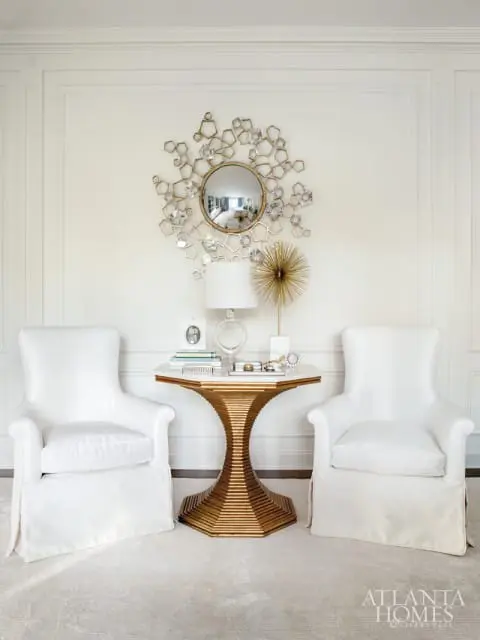 Two white slipcovered armchairs flank a gold pedestal table with a marble top, decorated with a sunburst mirror and a lamp.