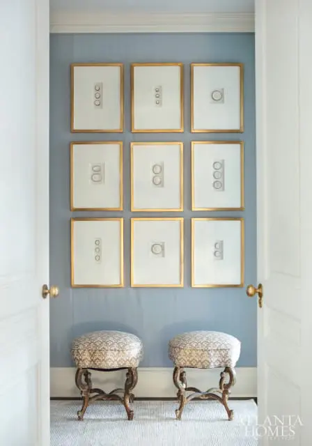 Hallway with pale blue walls, a grid of nine framed prints with gold frames, and two decorative stools with patterned upholstery.