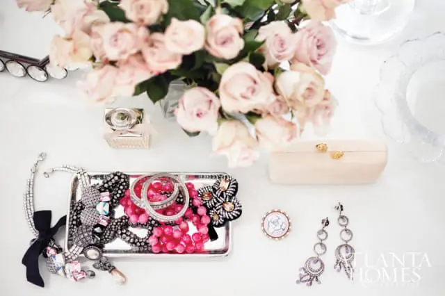 Silver tray filled with assorted jewelry including pink beaded necklaces, diamond bracelets, and a black ribbon bow. A bouquet of light pink roses sits behind the tray.
