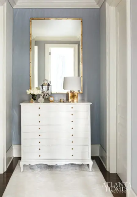 Hallway with pale blue textured walls, a white dresser with brass hardware, a gold-framed mirror, and a white rug on dark wood floors.