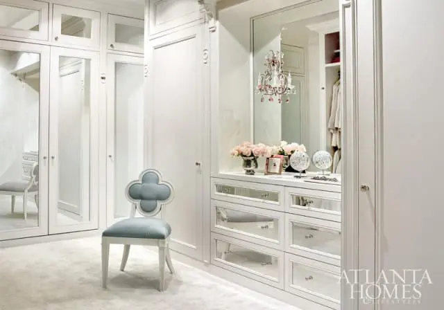White walk-in closet with mirrored drawers, a light blue quatrefoil chair, and a crystal chandelier reflecting in the mirror.