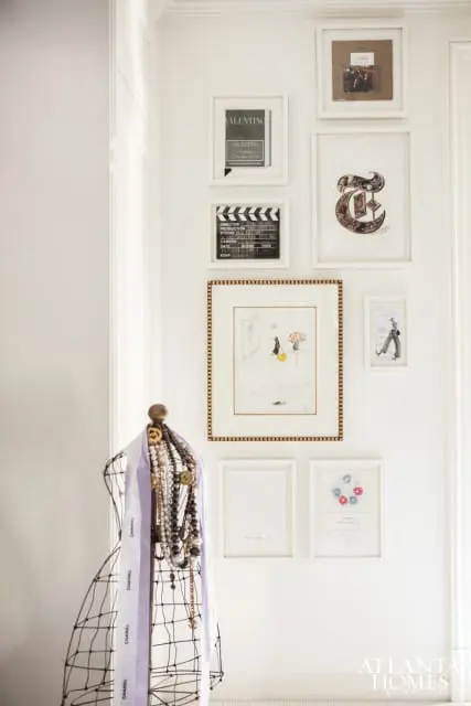 Wall with framed art including Valentino advertisements, a clapperboard, a fashion illustration, and a decorative letter 'T'. A wire mannequin displays necklaces and a Chanel scarf.
