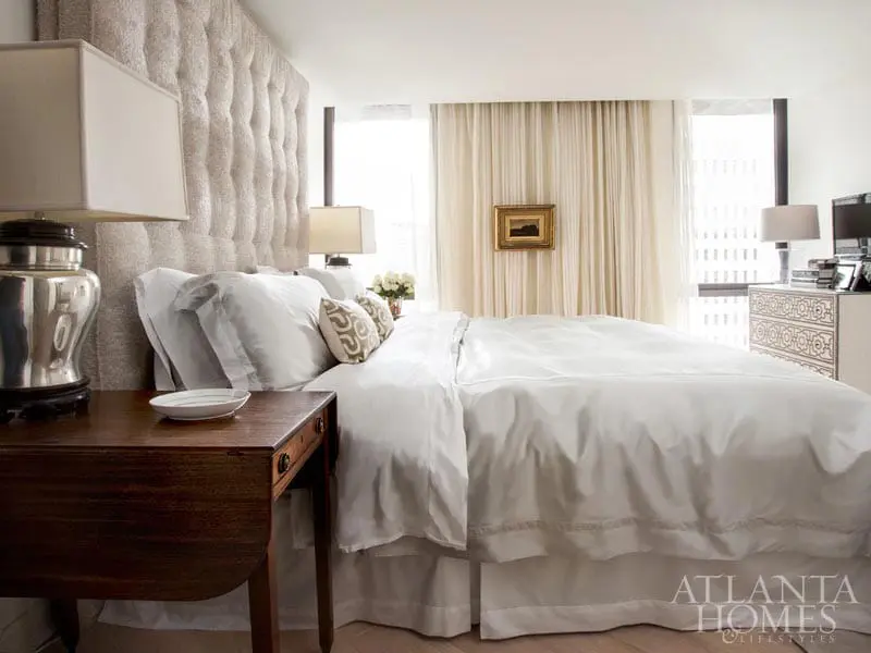 Bedroom with a tufted upholstered headboard, crisp white bedding, a dark wood nightstand with a silver lamp, and sheer curtains framing a city view.