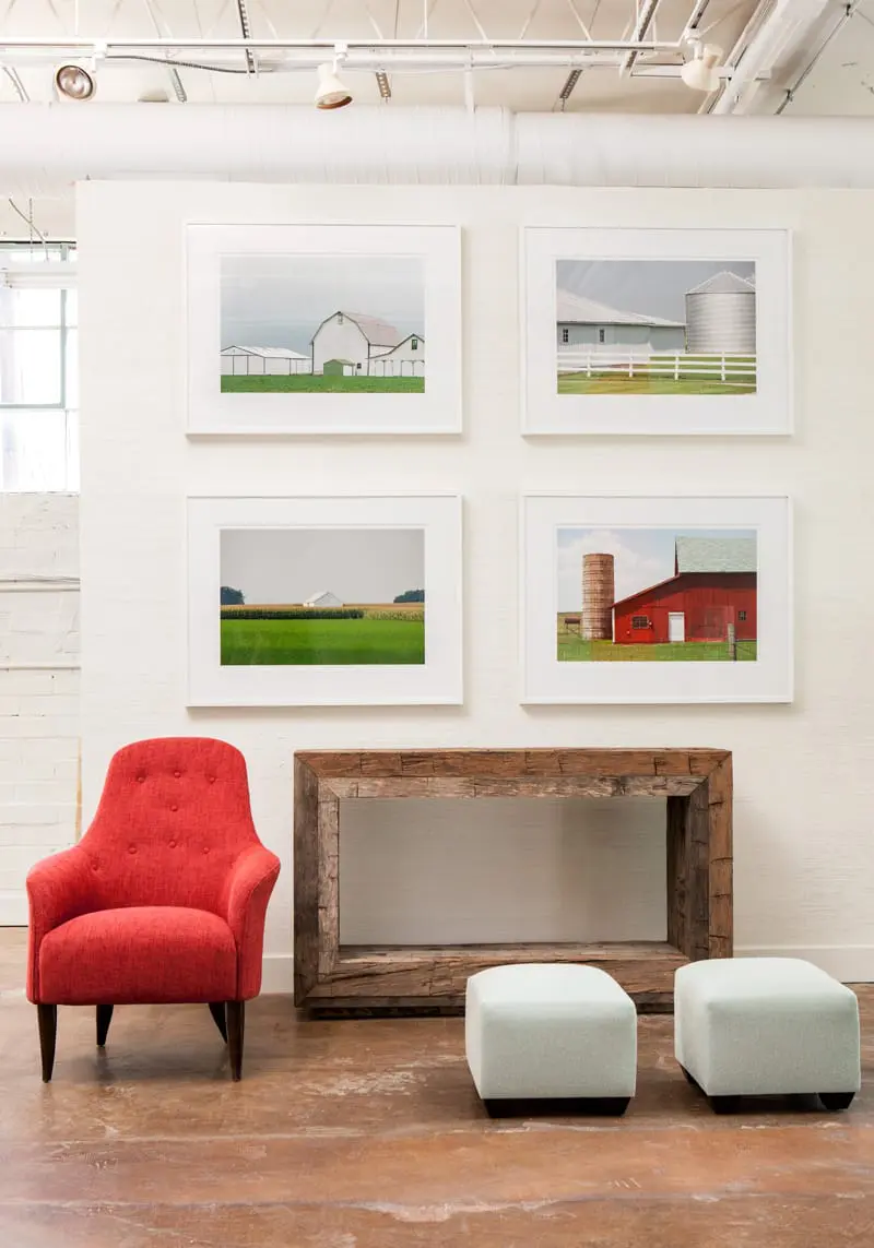 White brick wall with four framed photographs of barns and silos, a red armchair, a rustic wooden console table, and two light blue ottomans