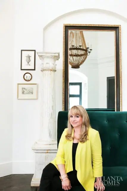 Woman with blonde hair wearing a yellow blazer and black top, sitting on a green velvet sofa in front of a large mirror and a decorative column.
