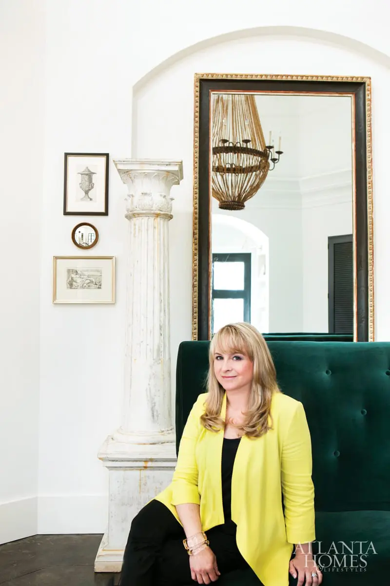 Woman with blonde hair wearing a yellow blazer and black top, sitting on a green velvet sofa in front of a large mirror and a decorative column.