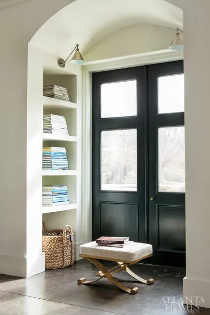 Entryway with built-in white shelves filled with books, a woven basket, a gold X-base ottoman with books, and dark green double doors with glass panes.