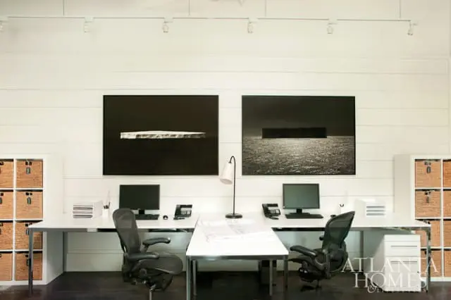 Modern office with white shiplap walls, two long white desks with black office chairs, computers, and two large black and white abstract art pieces depicting icebergs and water.