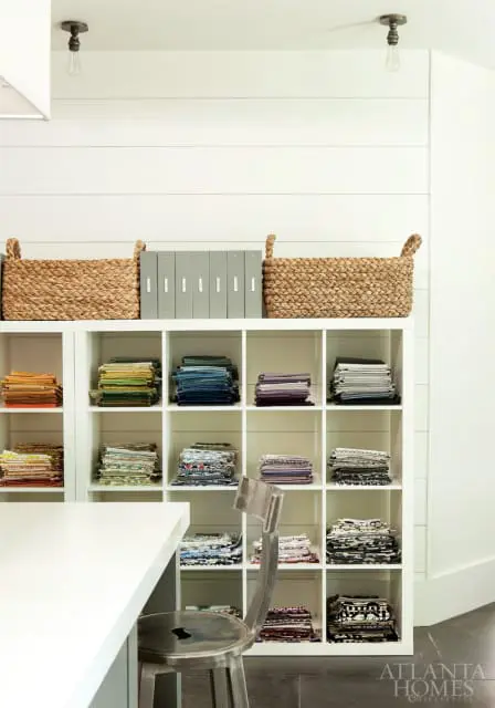 White shiplap wall with a white cube shelving unit filled with folded fabrics and woven baskets. A metal stool sits in front of the shelves.
