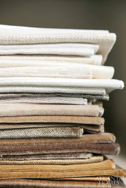 Stack of fabric swatches in neutral tones, from cream and beige to brown and gold, with varied textures including velvet and woven patterns.