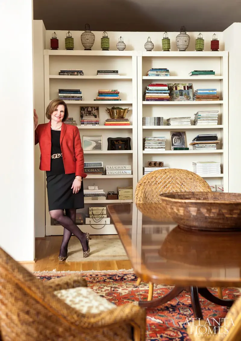 Woman in red blazer and black dress standing by white built-in bookshelves filled with books and decorative items.