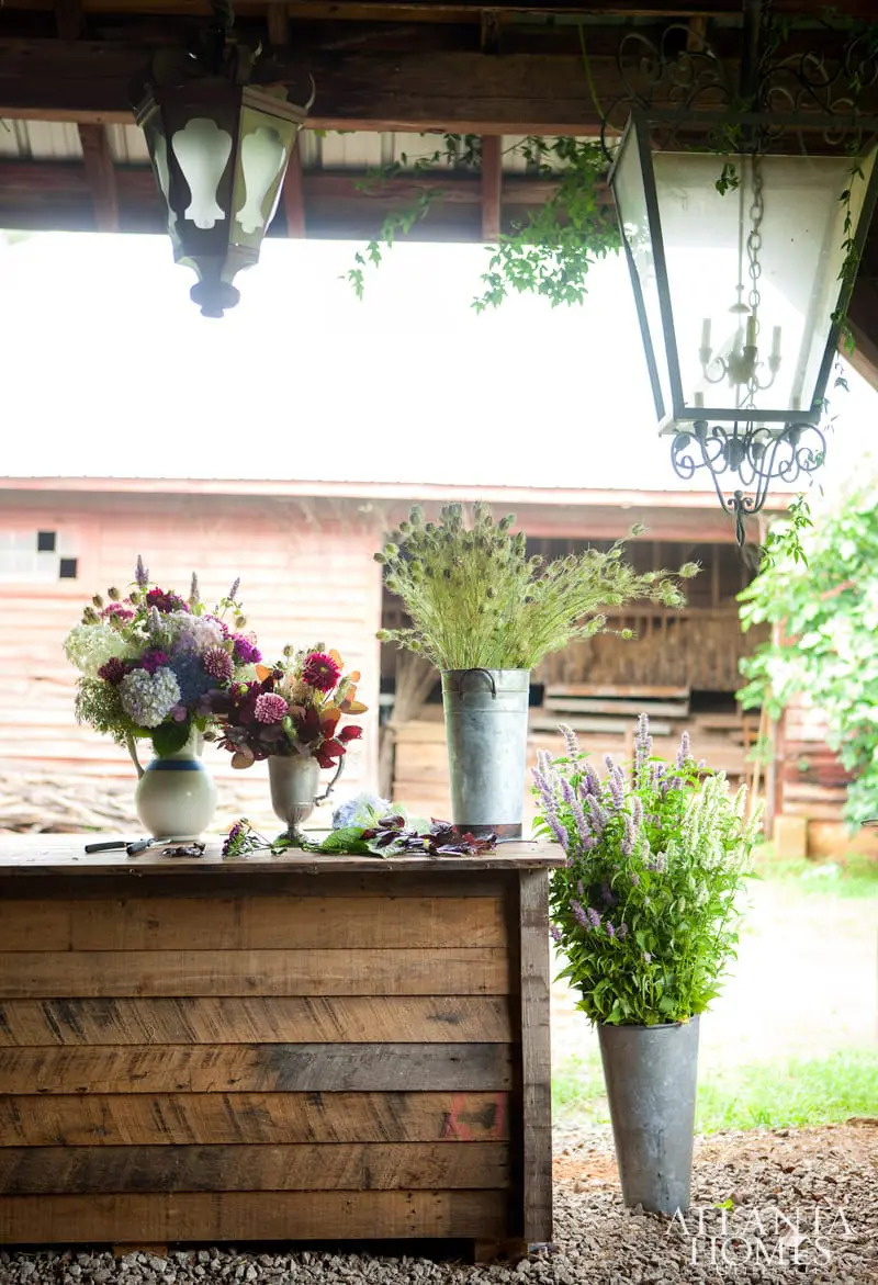 Wooden counter with floral arrangements in vases and buckets, under a covered porch with hanging lanterns.