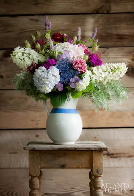 A bouquet of white, pink, purple, and blue flowers in a white pitcher with blue stripes, set on a rustic wooden table against a wooden plank background.