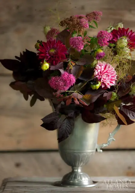 A bouquet of dark pink and burgundy dahlias, pink astilbe, and dark foliage in a silver urn against a wooden background.