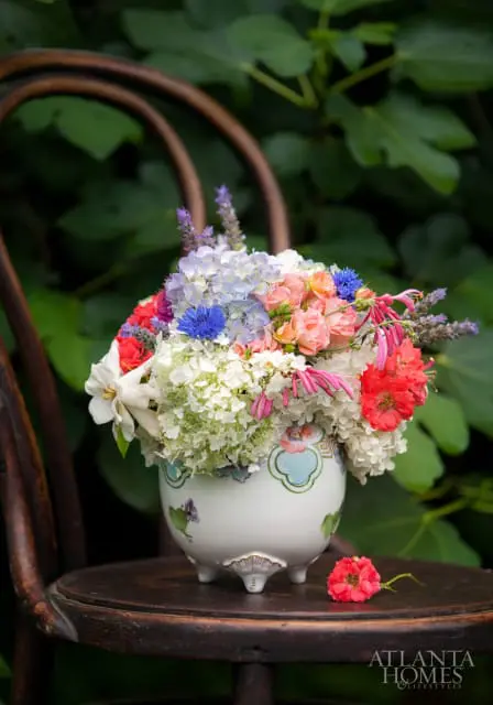 A colorful bouquet of hydrangeas, roses, lavender, and cornflowers in a decorative white vase with floral accents, resting on a dark wooden chair with lush green foliage in the background.
