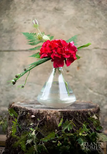 Red hydrangea and fern arrangement in a clear glass vase on a mossy tree stump.