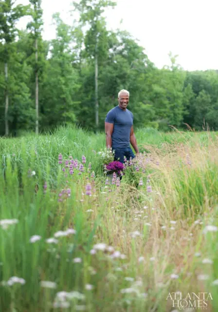 Man with blond hair wearing a blue t-shirt and jeans, holding a bucket of purple flowers, standing in a field of tall green grass and purple wildflowers.