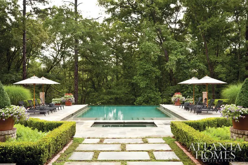 Rectangular swimming pool and spa surrounded by manicured hedges, flagstone patio, lounge chairs, umbrellas, and lush trees.