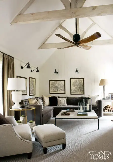 Living room with vaulted ceiling, exposed wooden beams, and a large wooden ceiling fan. Features a charcoal sectional sofa, upholstered armchairs, a white marble coffee table, and paneled walls adorned with botanical prints.