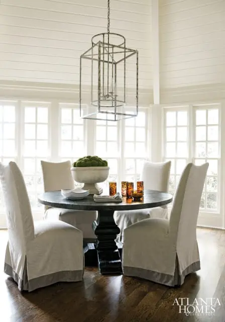 Dining room with dark pedestal table, four slipcovered chairs, large metal lantern chandelier, and a bowl of moss