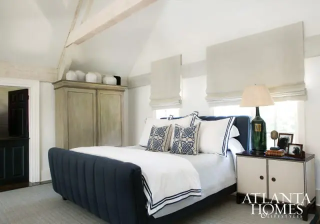 Bedroom with navy upholstered bed, white bedding with navy trim, patterned pillows, a distressed wood armoire, and a bedside table with a green lamp.