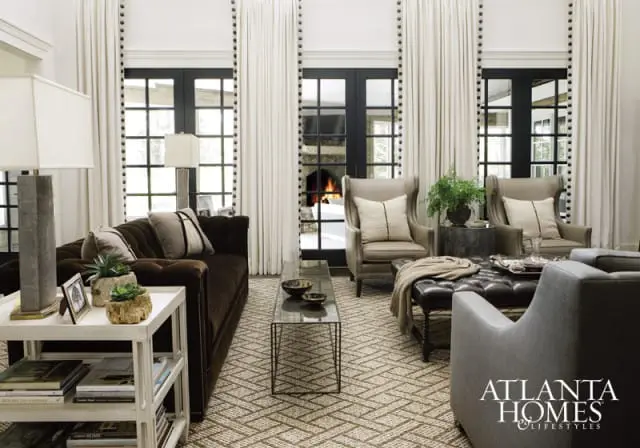 Living room with a dark brown tufted sofa, two taupe wingback chairs, a leather ottoman, and a glass coffee table in front of French doors with a fireplace visible beyond.