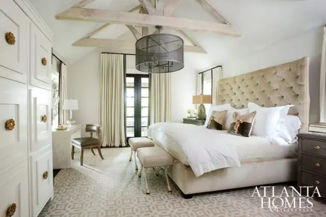 Bedroom with a tufted beige headboard, white bedding, and a large white wardrobe with gold hardware. Exposed wooden beams accent the vaulted ceiling.