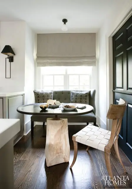 Breakfast nook with dark wood floors, a gray banquette, round metal table with cheese and olives, and a wooden chair with white woven seat.