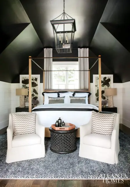 Bedroom with a black geometric ceiling, four-poster wood bed with white bedding, two white armchairs, and a dark patterned rug.