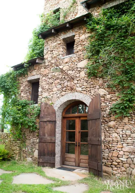 Stone building with arched wooden double doors, wooden shutters, and ivy growing on the facade.