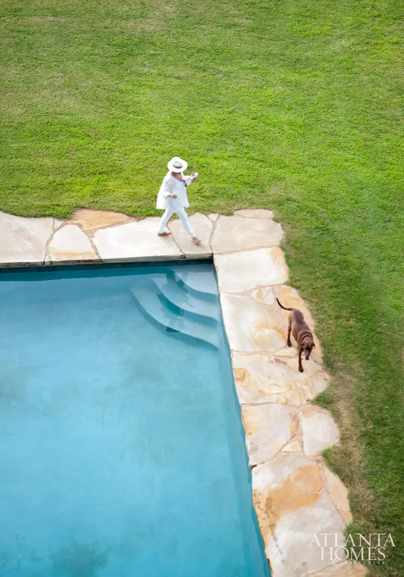 Woman in white walking by a blue swimming pool with a dog on flagstone patio and green grass