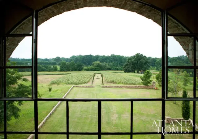 View through arched window of a formal garden with a central pathway leading to a fountain, flanked by sunflower fields and surrounded by trees.