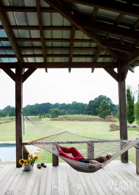 Person relaxing in a hammock on a wooden porch overlooking a field of sunflowers and trees.