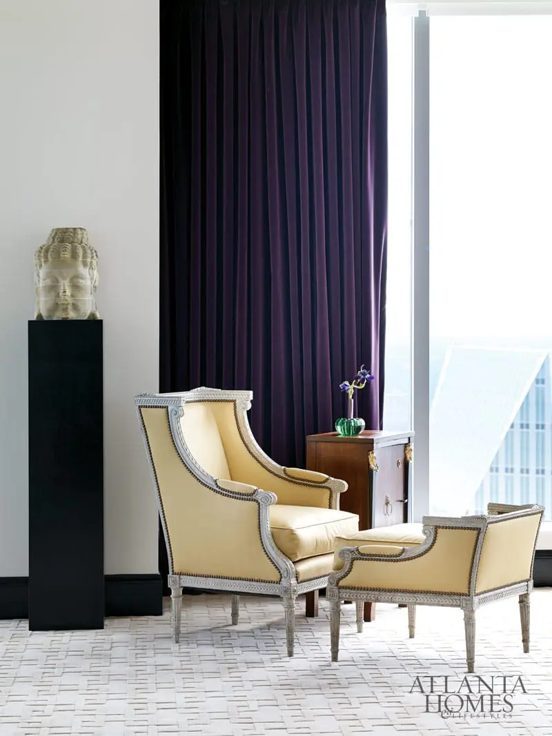 Two cream upholstered armchairs with ornate carved wood frames sit on a cream and white geometric rug. A dark wood cabinet with brass hardware holds a green glass vase with purple flowers. A stone Buddha head sculpture on a black pedestal stands against a white wall.