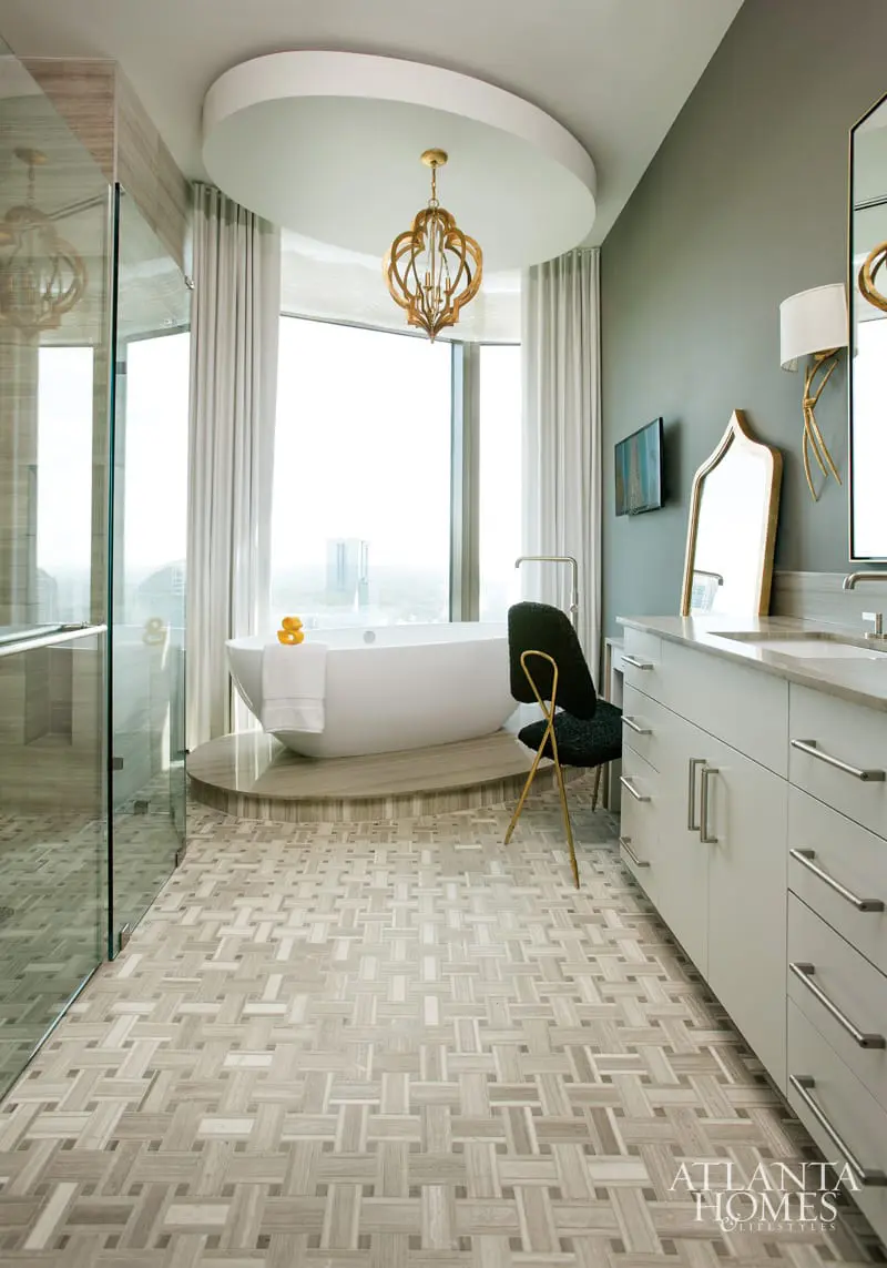Modern bathroom with freestanding tub, glass shower, double vanity, and intricate wood-look tile flooring. A gold chandelier hangs from a curved ceiling.