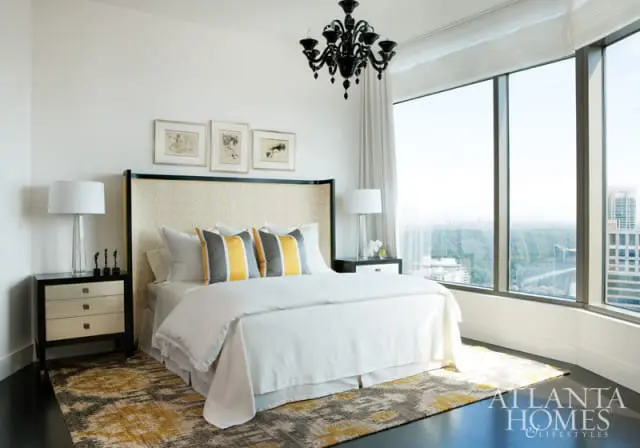 Bedroom with a white bed, yellow and gray striped pillows, a patterned headboard, a black chandelier, and a large window with a city view.